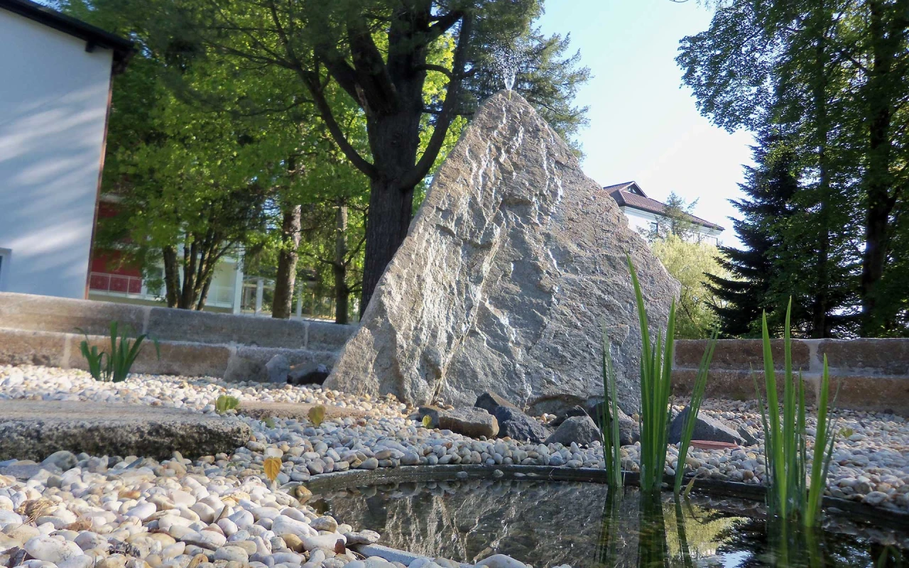 Bild Stein KModerner Zierbrunnen mit großem Naturstein, Kiesbeet und Wasserlauf. Die harmonisch gestaltete Gartenanlage lädt zum Verweilen und Entspannen ein.linikpark