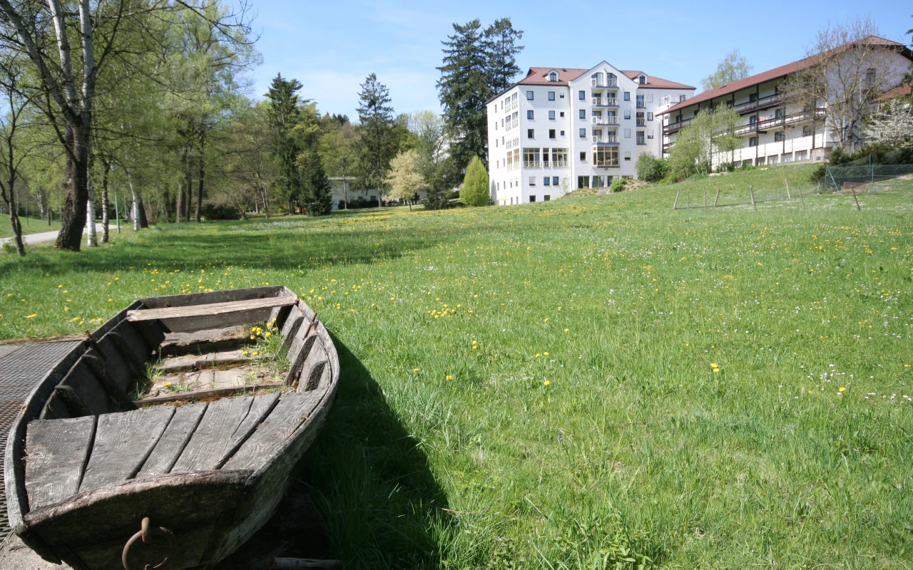 Bild Liegewiese Blick auf das Klinikgebäude mit weitläufiger Grünfläche, altem Ruderboot und schattenspendenden Bäumen im Vordergrund.